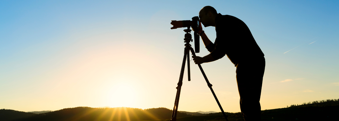 The silhouette of a man in front of a sunset, taking photos using a camera set up on a tripod. 