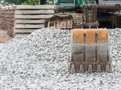 the bucket of a backhoe resting on some gravel with stacks of brick in the background