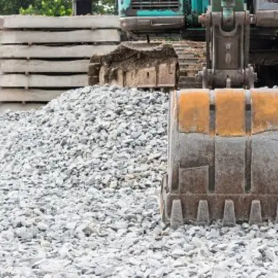the bucket of a backhoe resting on some gravel with stacks of brick in the background