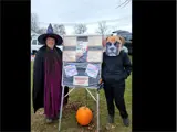 Woman to the left dressed up as a witch and inidvidual on the right dressed up as a dog standing in front of a bulletin board.