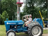 Man sitting on a blue Ford tractor watering a floral hanging basket.
