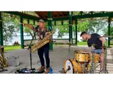 Two men playing instruments. The man on the left is playing saxophone and the man on the right is playing drums.